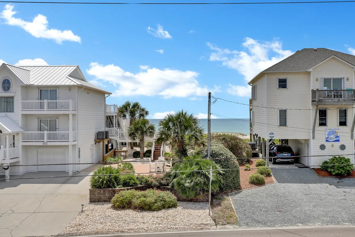 A picturesque exterior view of the beach, flanked by nearby palm trees and two neighboring beach houses. The scene captures inviting natural light and the proximity to the ocean, evoking early morning walks and sunset picnics.