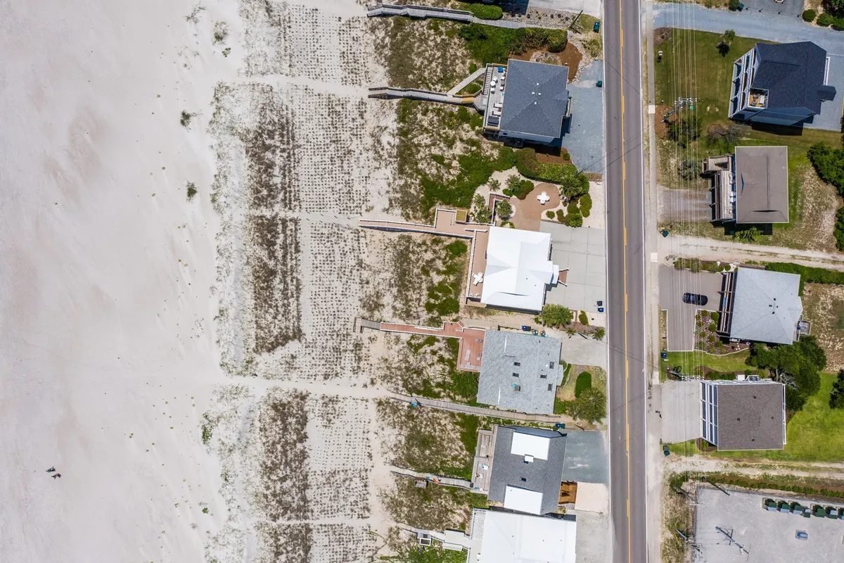 A casual, inviting exterior scene showing beachfront properties and a sandy dune just across a road, with the ocean visible beyond. The image captures people enjoying the beach, walking along the shore, and gathering in small groups, evoking a vibrant beachside community atmosphere.