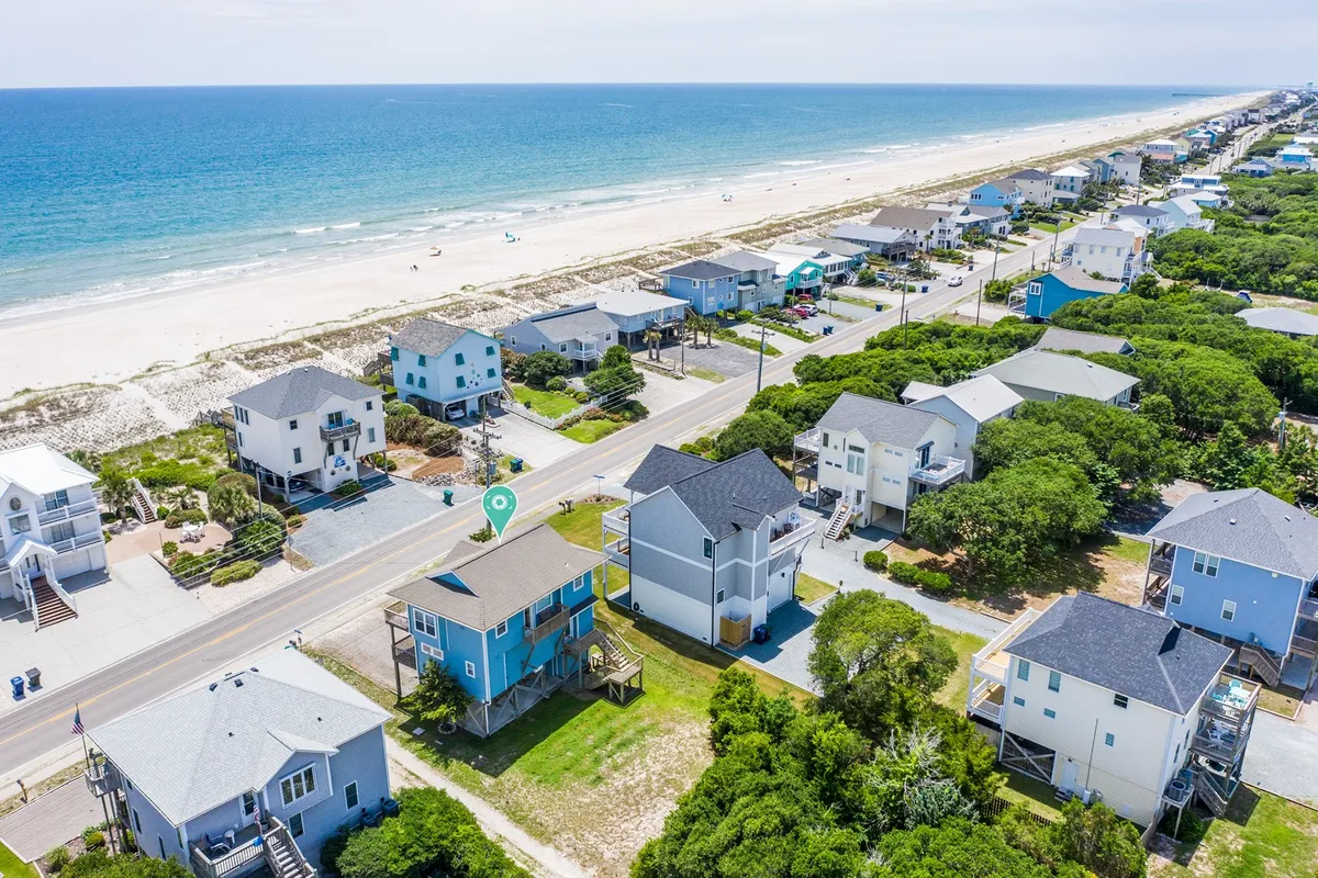 A cheerful exterior shot capturing colorful beachfront houses, a vibrant green landscape, and a clear view of the beach and ocean. The setting is lively, with a road separating the homes from the sand, hinting at easy access to both beach activities and neighborhood amenities.