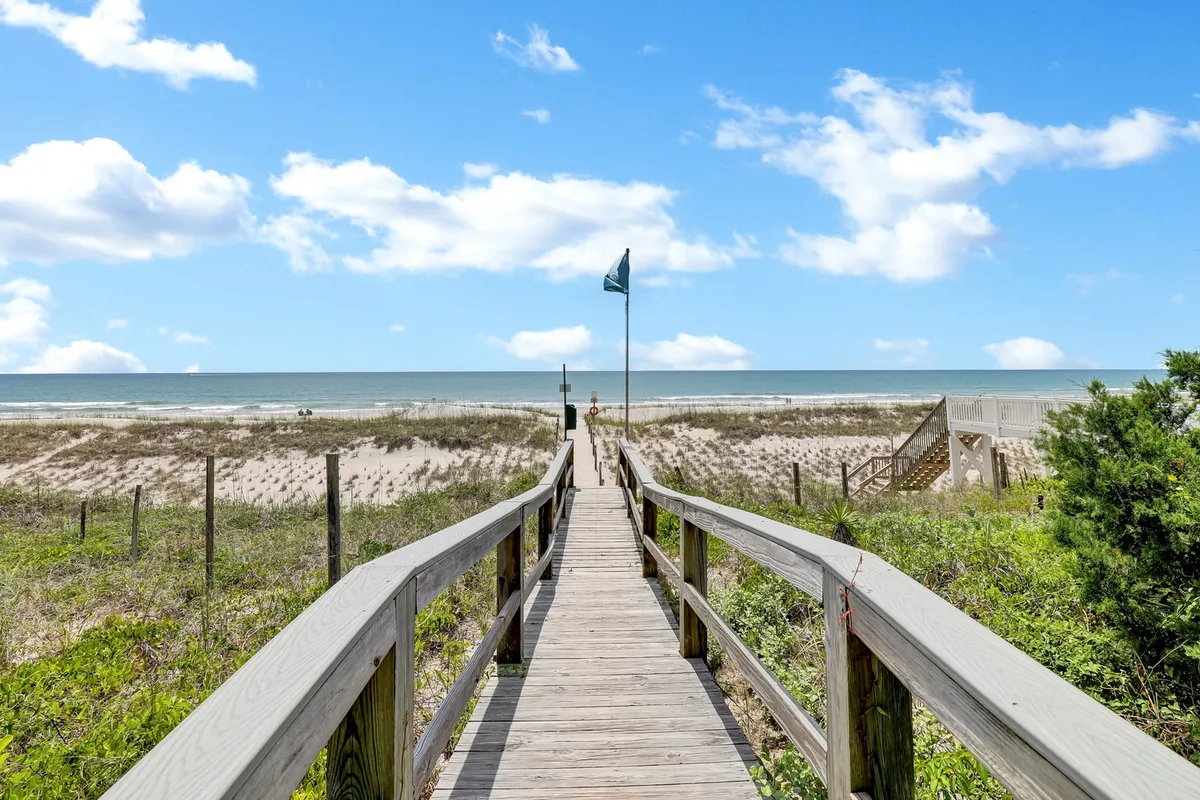 A peaceful wooden boardwalk extends through dune grasses, leading directly to the sandy shore and the blue ocean beyond. The scene is sunlit, inviting, and captures the anticipation of a day at the beach.