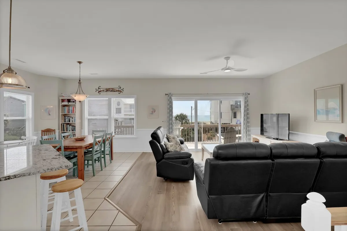 The image captures a cozy living area with a comfortable leather sofa, a dining table with colorful chairs, and large sliding glass doors opening to the outdoors. The space is filled with natural light and provides clear visual access to the ocean beyond, enhancing the sense of being surrounded by coastal beauty.