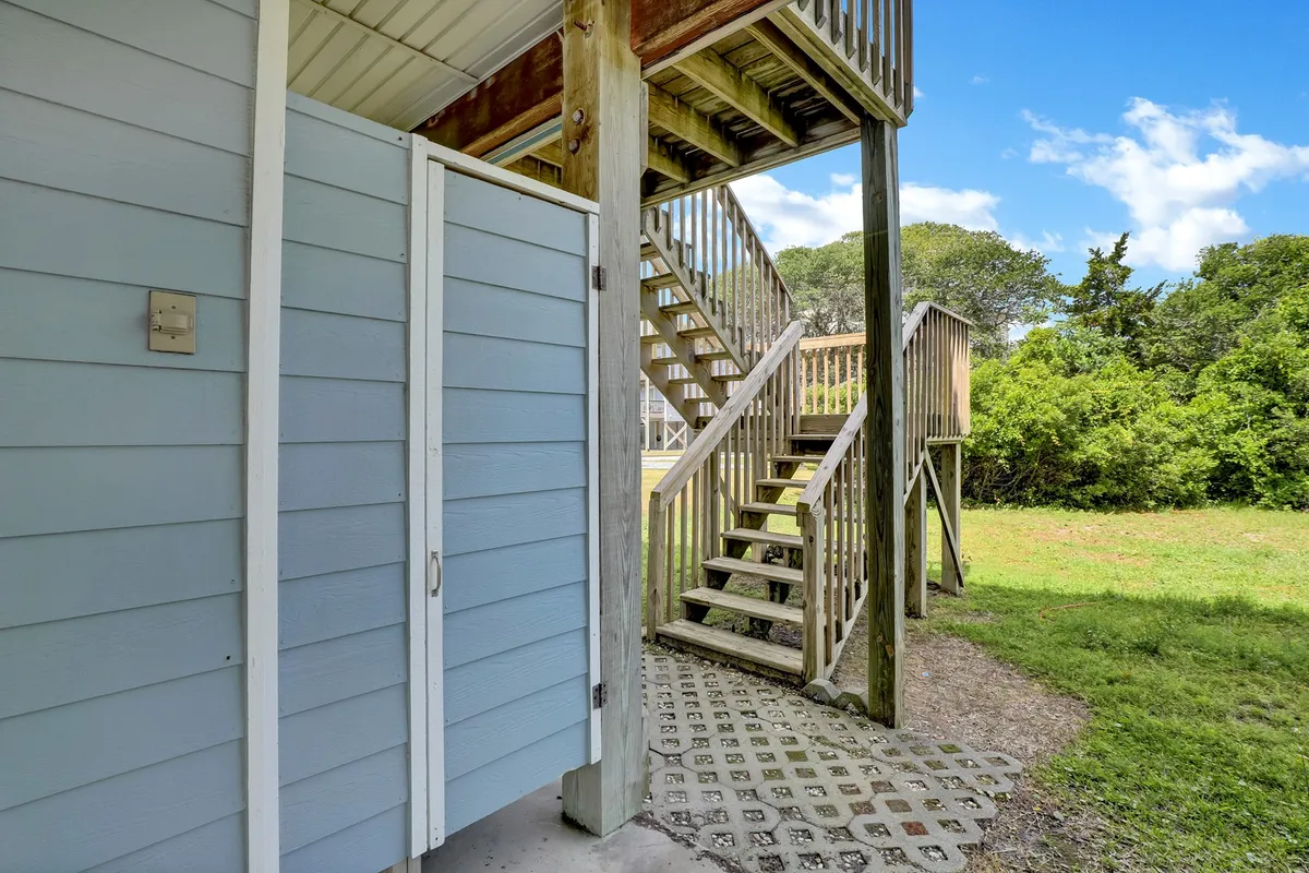 This image captures an exterior view of the house with a visible outdoor shower installed next to a blue-sided wall and staircase. The area around the shower is open and relaxed, suggesting easy access for rinsing off after the beach. The yard space is inviting, hinting at family gatherings or transitions from outdoor fun to indoor comfort.