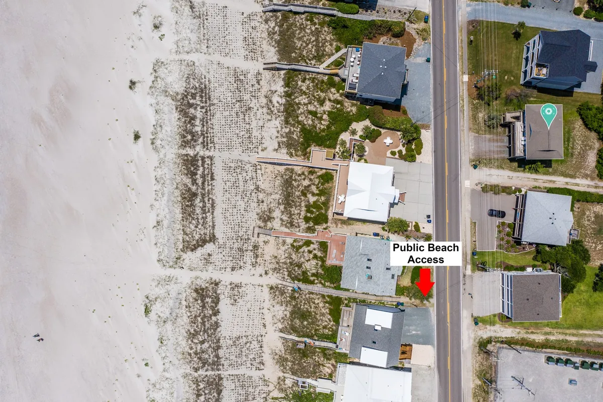 A relaxed exterior scene featuring a public beach access sign, nearby coastal houses, and sand dunes leading toward the beach. The pathway to the beach is visually clear, emphasizing how close and effortless beach access is from the home.