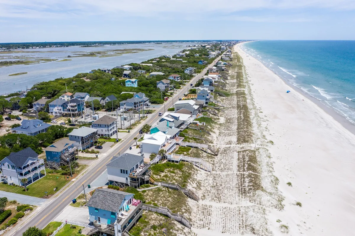 An expansive exterior shot showcasing beachfront houses, a coastal road ideal for biking, a wide sandy beach, gentle ocean waves, and surrounding marshland. The scene highlights the relaxed, outdoor lifestyle and the natural landscapes waiting to be explored.