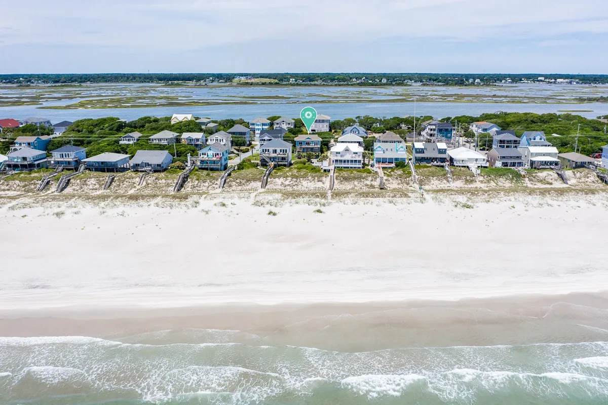 This image showcases a row of beachfront houses with sand dunes, gentle ocean waves, and a green marker indicating the property's location. The road between the homes and the beach is clearly visible, emphasizing immediate access to the shore.