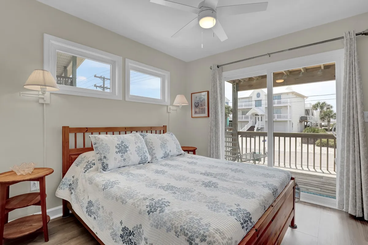 A tranquil bedroom with floral bedding on a large bed, large windows letting in natural light, and a wooden bedside table, suggesting a serene morning experience with views of the outdoors.