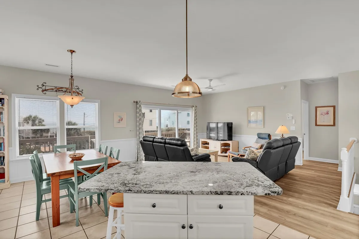 This image features an open-concept space combining a granite kitchen island, dining table, and a comfortable seating area. Large windows flood the room with natural light, and the layout is oriented to showcase the ocean views just beyond. The bright, airy atmosphere invites relaxation and socializing, perfectly matching the section's emphasis on gathering spaces.